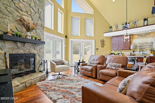 living area featuring french doors, a towering ceiling, wood finished floors, a stone fireplace, and a notable chandelier