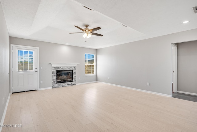 unfurnished living room featuring baseboards, light wood-style flooring, ceiling fan, a tray ceiling, and a stone fireplace