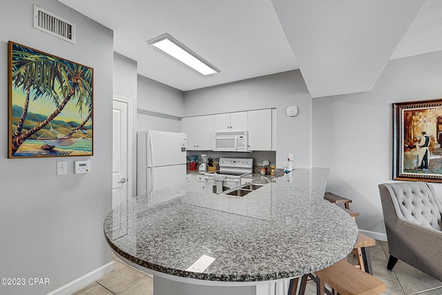 kitchen with a peninsula, white appliances, visible vents, a kitchen breakfast bar, and dark stone countertops