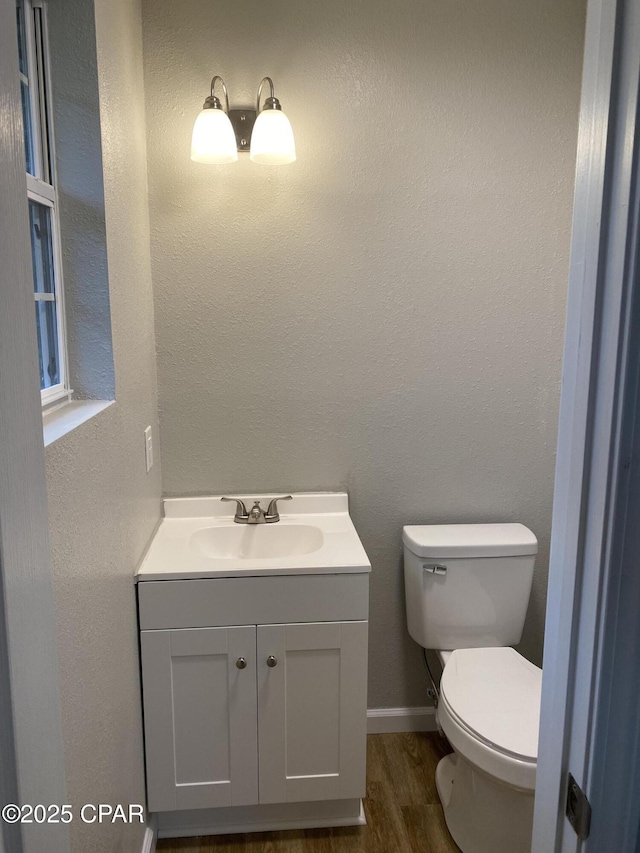 half bath featuring vanity, wood finished floors, baseboards, toilet, and a textured wall