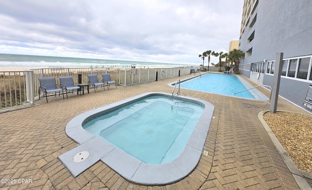 community pool with a patio area, a beach view, fence, and a water view