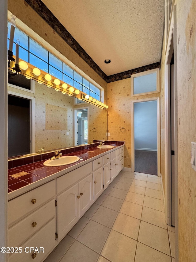 full bathroom with double vanity, a textured ceiling, wallpapered walls, and a sink