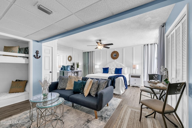 bedroom featuring a ceiling fan, wood finished floors, visible vents, a paneled ceiling, and a decorative wall