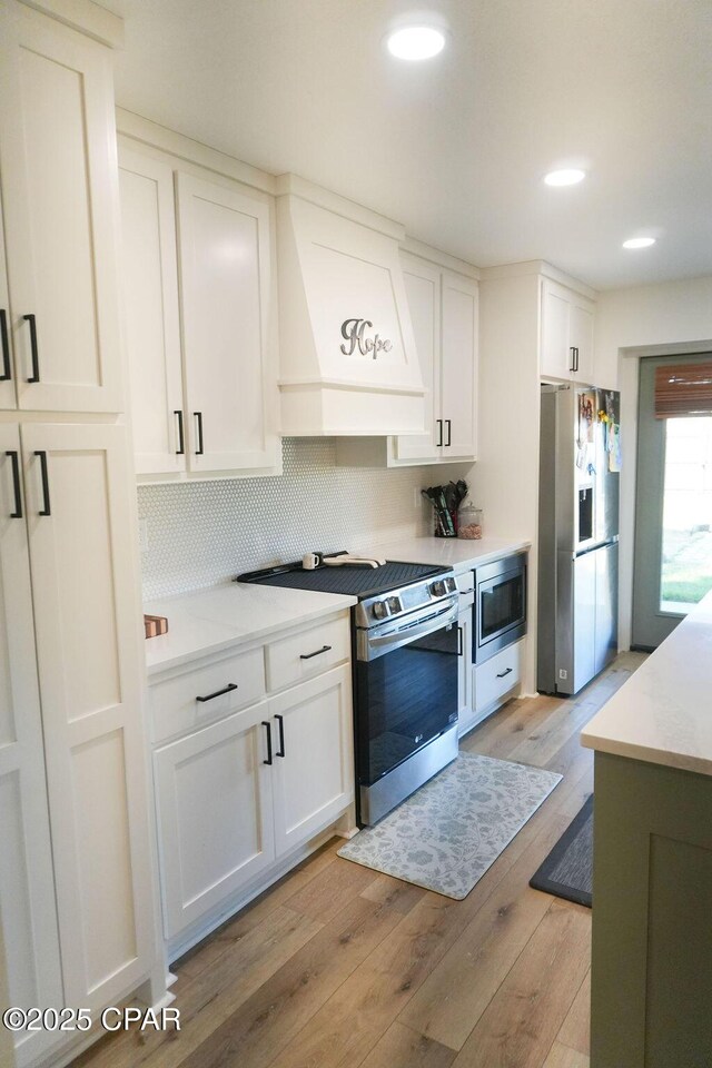 kitchen with custom exhaust hood, light wood-style flooring, decorative backsplash, white cabinets, and appliances with stainless steel finishes