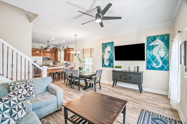 living room with ceiling fan with notable chandelier, light wood-style floors, crown molding, baseboards, and stairs