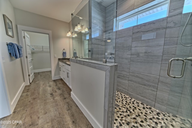 bathroom featuring wood finished floors, tiled shower, vanity, baseboards, and a spacious closet