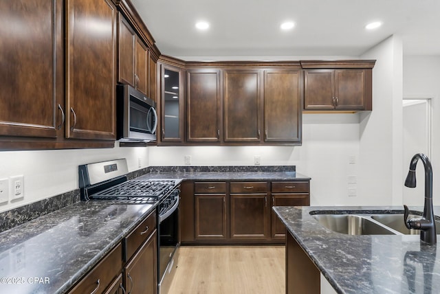 kitchen with light wood finished floors, dark brown cabinetry, dark stone counters, appliances with stainless steel finishes, and a sink