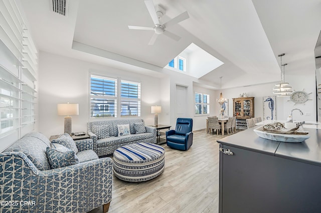 living room featuring visible vents, ceiling fan with notable chandelier, a tray ceiling, light wood finished floors, and lofted ceiling