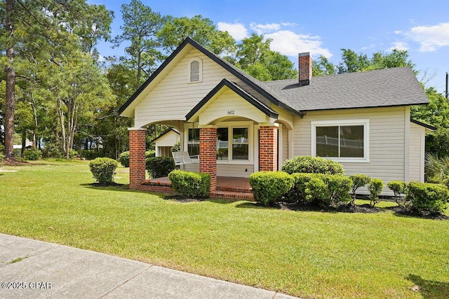view of front facade with roof with shingles, a front yard, brick siding, a porch, and a chimney