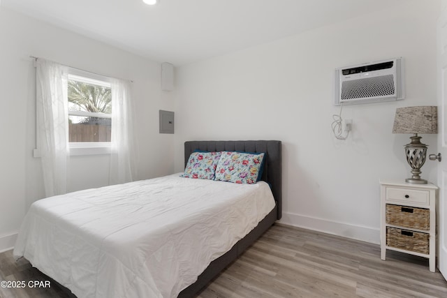 bedroom featuring wood finished floors, baseboards, a wall mounted AC, and electric panel
