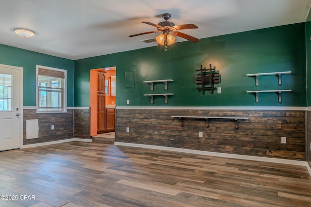 unfurnished living room featuring wooden walls, wainscoting, visible vents, ceiling fan, and wood finished floors
