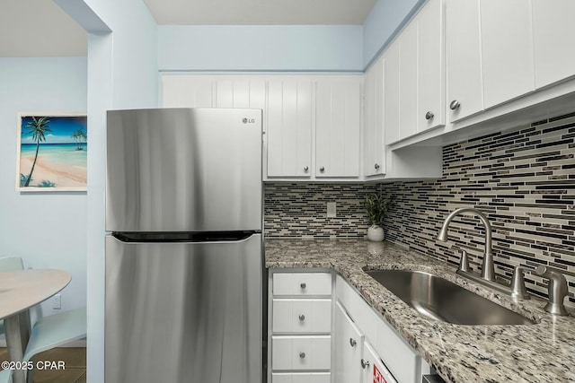 kitchen featuring white cabinets, a sink, freestanding refrigerator, decorative backsplash, and light stone counters