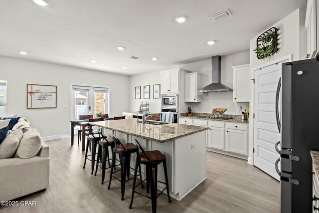kitchen featuring wall chimney range hood, appliances with stainless steel finishes, light stone counters, open floor plan, and visible vents