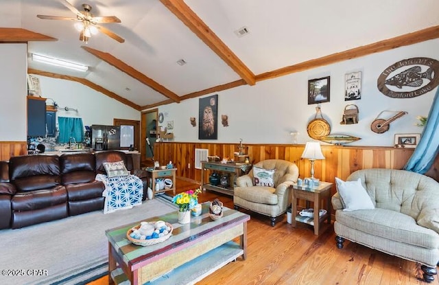living room with lofted ceiling with beams, wainscoting, light wood-type flooring, ceiling fan, and wooden walls
