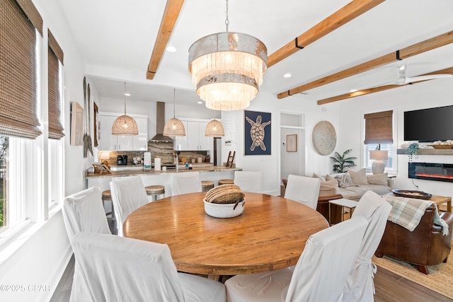 dining area featuring a glass covered fireplace, wood finished floors, beam ceiling, recessed lighting, and ceiling fan with notable chandelier