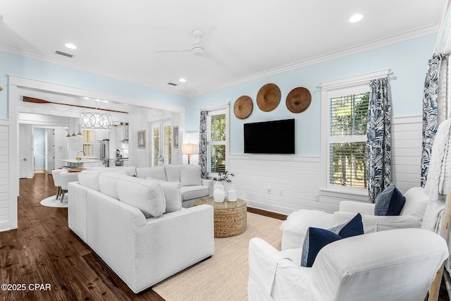 living area with a wainscoted wall, dark wood-style flooring, visible vents, ceiling fan with notable chandelier, and ornamental molding