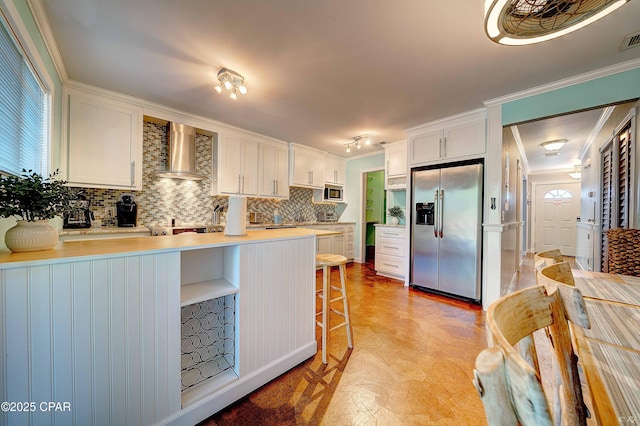 kitchen featuring appliances with stainless steel finishes, wall chimney exhaust hood, a peninsula, white cabinetry, and crown molding