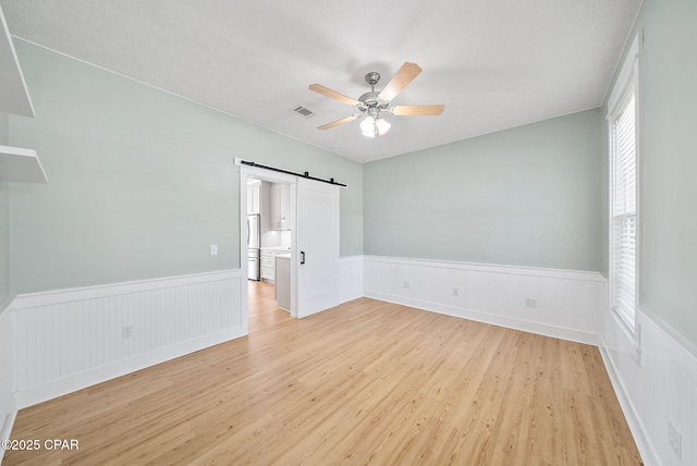 unfurnished bedroom featuring a barn door, light wood finished floors, ceiling fan, visible vents, and a wainscoted wall