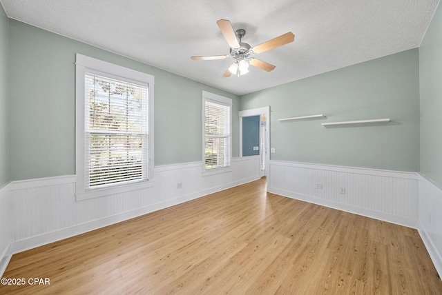 unfurnished room with a ceiling fan, a textured ceiling, light wood-style flooring, and a wainscoted wall