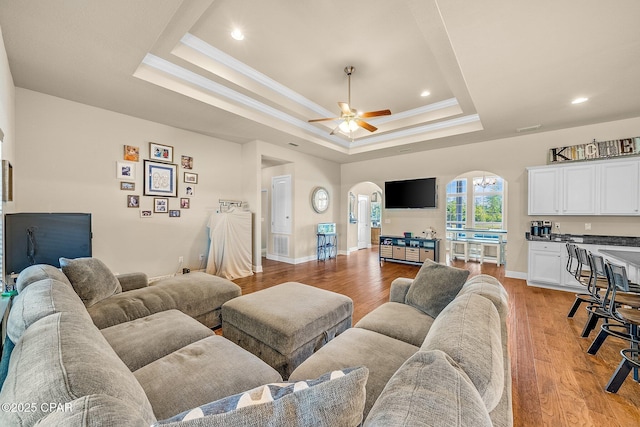 living area with arched walkways, a tray ceiling, a ceiling fan, and light wood-style floors