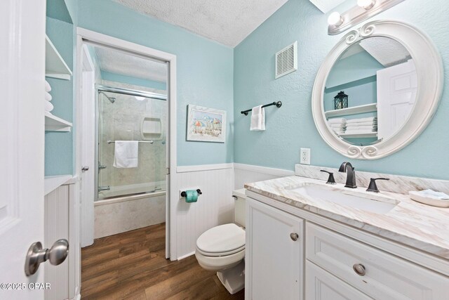 8743 Thomas Dr Unit 809, Panama City Beach, FL 32408 - bathroom with a textured ceiling, wood finished floors, toilet, a wainscoted wall, and combined bath / shower with glass door