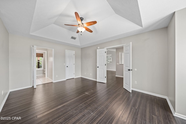unfurnished bedroom with a ceiling fan, baseboards, dark wood-type flooring, a tray ceiling, and visible vents