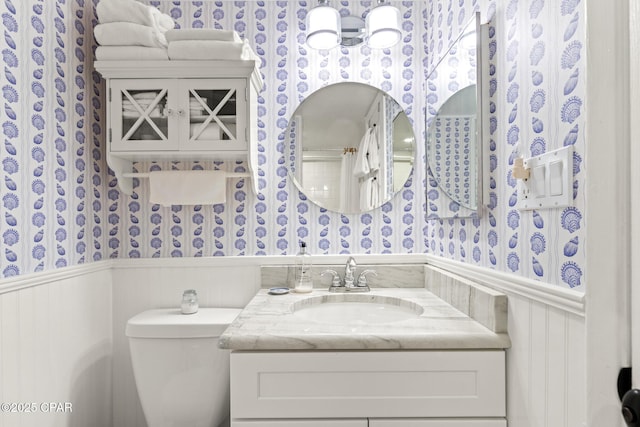 bathroom featuring vanity, toilet, wallpapered walls, and a wainscoted wall