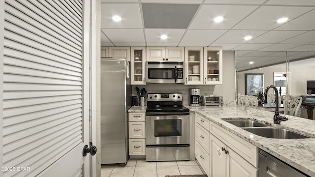 kitchen with stainless steel appliances, a sink, light tile patterned floors, light stone countertops, and glass insert cabinets