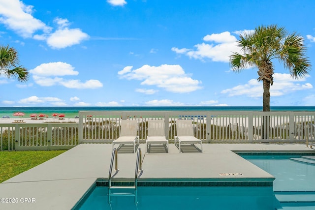 community pool with a patio, view of water and beach, and fence