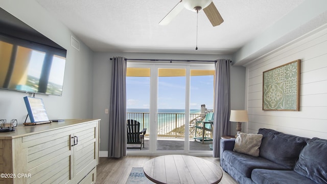 living room with view of water and beach, light wood-type flooring, a ceiling fan, and a textured ceiling