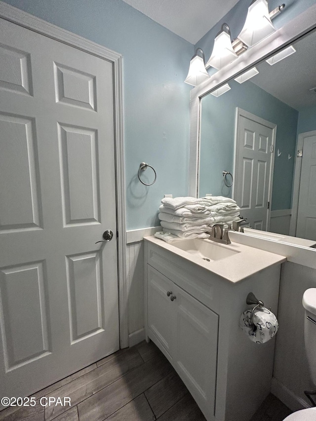 half bath featuring wood finished floors, toilet, vanity, and wainscoting