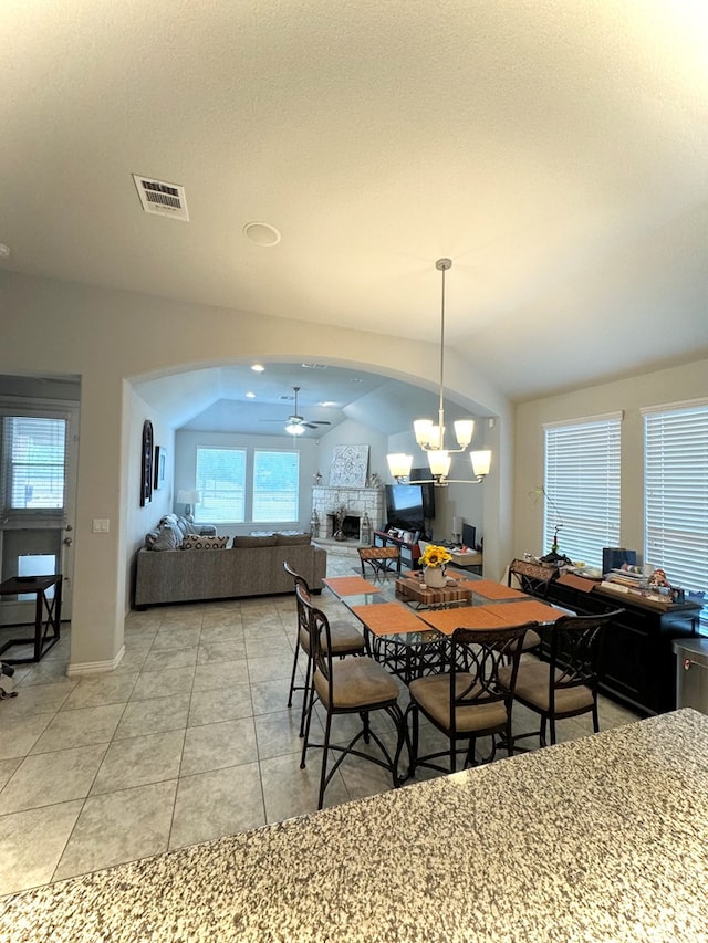 tiled dining space with ceiling fan with notable chandelier, vaulted ceiling, and a stone fireplace