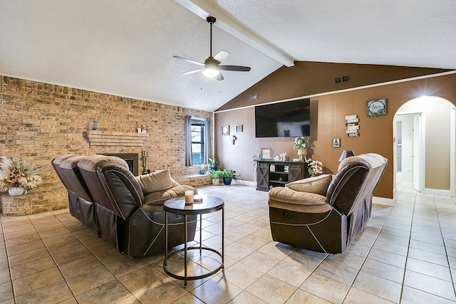 tiled living room with wood walls, vaulted ceiling with beams, a brick fireplace, a textured ceiling, and ceiling fan