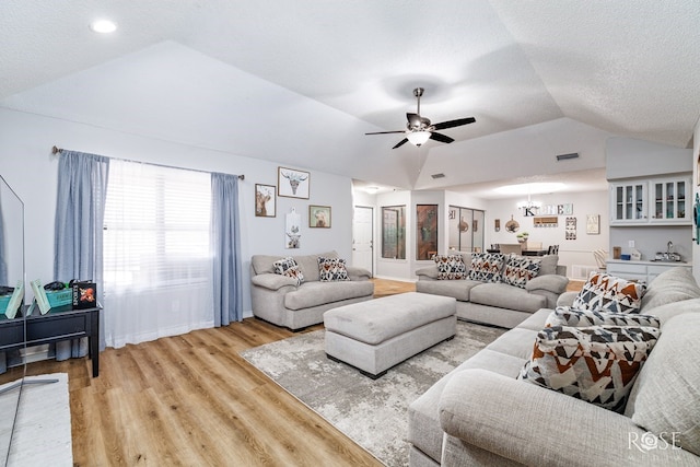 living room featuring visible vents, a textured ceiling, light wood finished floors, ceiling fan with notable chandelier, and vaulted ceiling