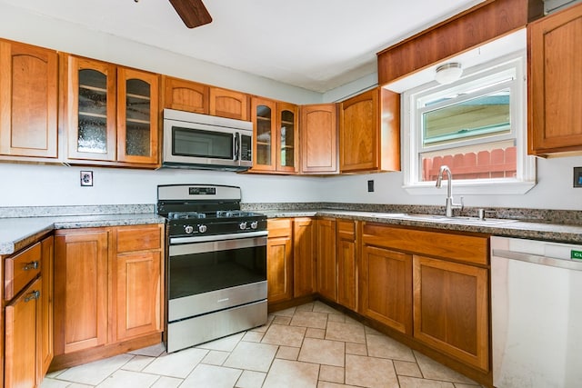 kitchen with brown cabinetry, a ceiling fan, glass insert cabinets, a sink, and stainless steel appliances