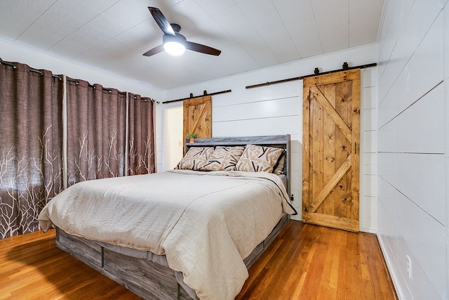 bedroom featuring wooden walls, hardwood / wood-style flooring, a ceiling fan, and a barn door