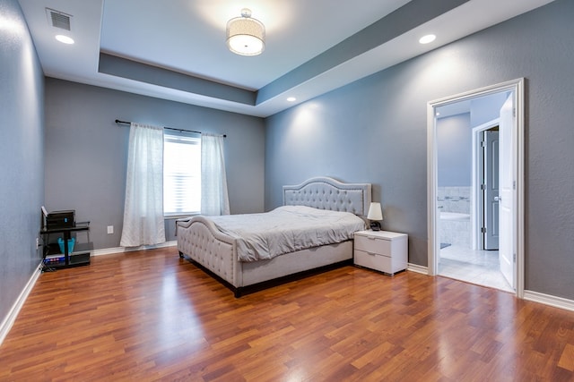bedroom featuring wood finished floors, baseboards, ensuite bathroom, a tray ceiling, and recessed lighting