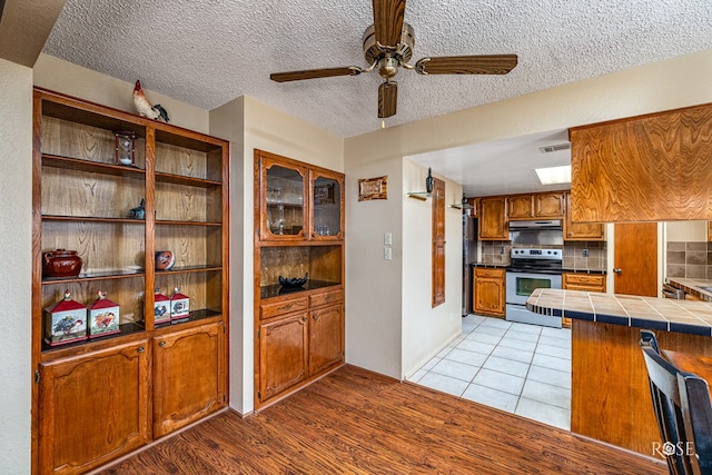 1729 Parkview Dr, San Angelo, TX 76904 - kitchen with stainless steel electric range, brown cabinets, a ceiling fan, under cabinet range hood, and tile countertops