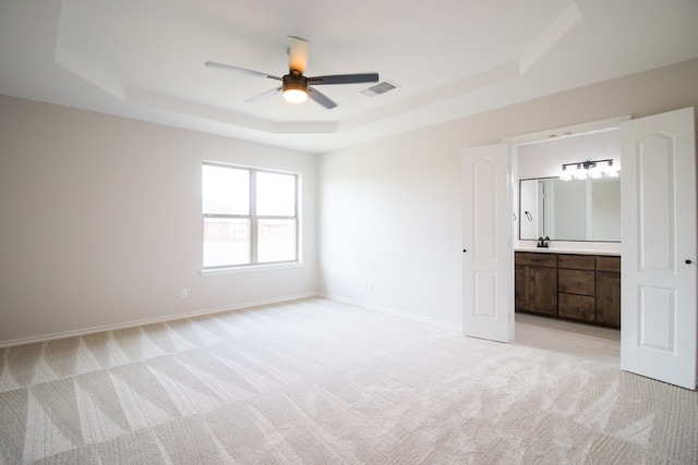 unfurnished bedroom with ceiling fan, ensuite bathroom, a raised ceiling, and light carpet