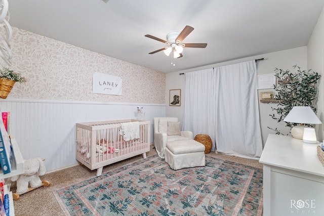 bedroom featuring a wainscoted wall, a nursery area, wallpapered walls, carpet floors, and ceiling fan