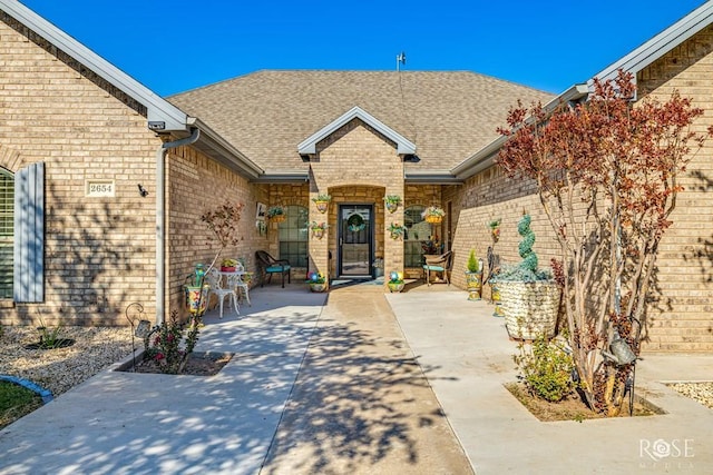 doorway to property featuring a shingled roof, brick siding, and a patio