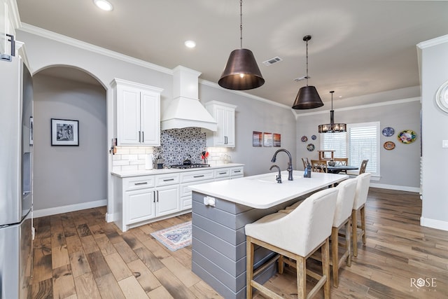 kitchen featuring an island with sink, white cabinets, hanging light fixtures, stainless steel appliances, and custom range hood