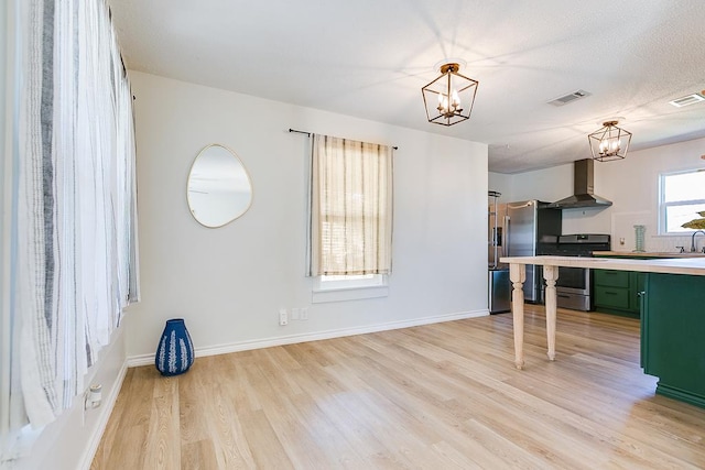 kitchen with light hardwood / wood-style flooring, appliances with stainless steel finishes, green cabinets, a notable chandelier, and wall chimney range hood