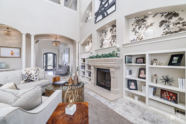 living room with arched walkways, a fireplace with raised hearth, built in shelves, a high ceiling, and ornate columns