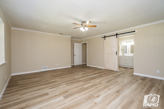 unfurnished bedroom featuring ensuite bathroom, crown molding, light wood-type flooring, ceiling fan, and a barn door