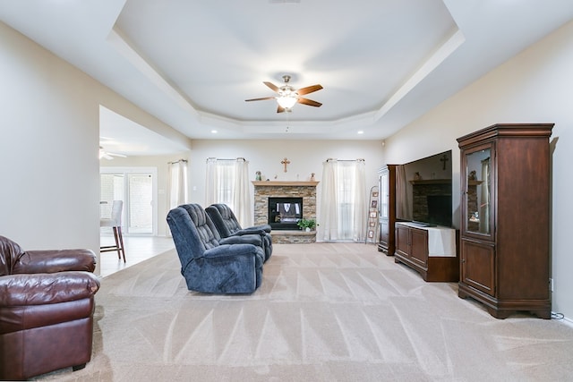 living area with a ceiling fan, a raised ceiling, light colored carpet, and a stone fireplace