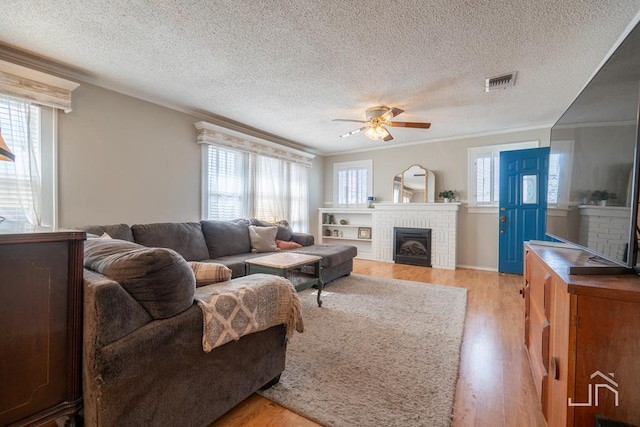 living room featuring visible vents, light wood finished floors, ornamental molding, a brick fireplace, and a wealth of natural light