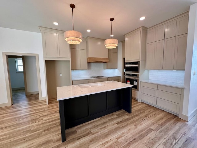 kitchen featuring light stone countertops, decorative light fixtures, light wood-style flooring, appliances with stainless steel finishes, and a kitchen island with sink