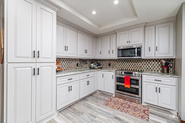 kitchen featuring light hardwood / wood-style flooring, stainless steel appliances, light stone counters, a tray ceiling, and decorative backsplash