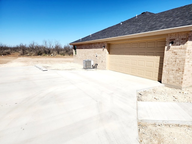 view of property exterior with driveway, a garage, roof with shingles, central air condition unit, and brick siding